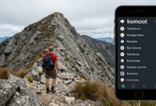 Um aventureiro no cume de uma montanha ao amanhecer, consultando um percurso de trilha em seu smartphone, que exibe a interface do aplicativo komoot.