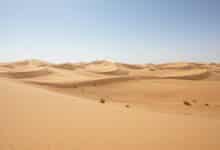 Uma vista aérea cinematográfica de um deserto vasto com dunas de areia onduladas sob um céu claro de final de tarde, destacando a textura geológica da paisagem.