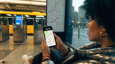 Pessoa consultando o aplicativo Moovit em um celular dentro de uma estação de transporte público, com ônibus e linhas de metrô representados em um mapa urbano.
