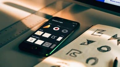 Celular exibindo uma interface de criação de logotipo com opções de ícones, fontes e paletas de cores ao lado de esboços de identidade visual em uma mesa de trabalho iluminada.