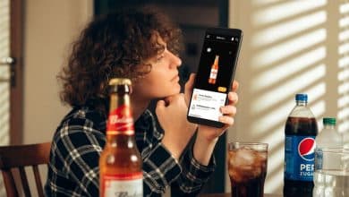 Pessoa consultando um aplicativo de entrega de bebidas no celular em casa, com cervejas, refrigerantes e água sobre uma mesa.